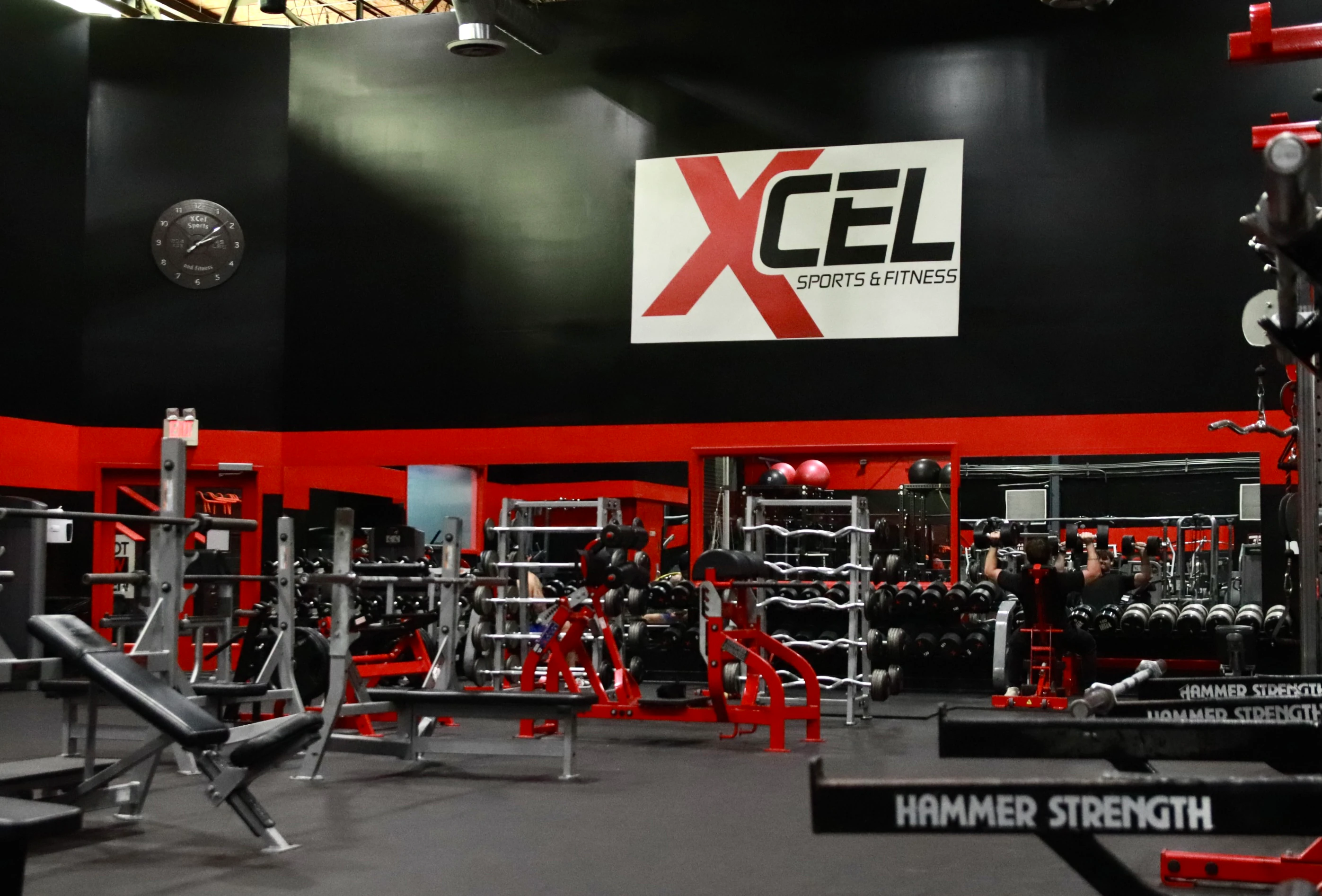 XCEL Sports & Fitness sign above Hammer Strength racks and benches at the gym in Fairfield, NJ