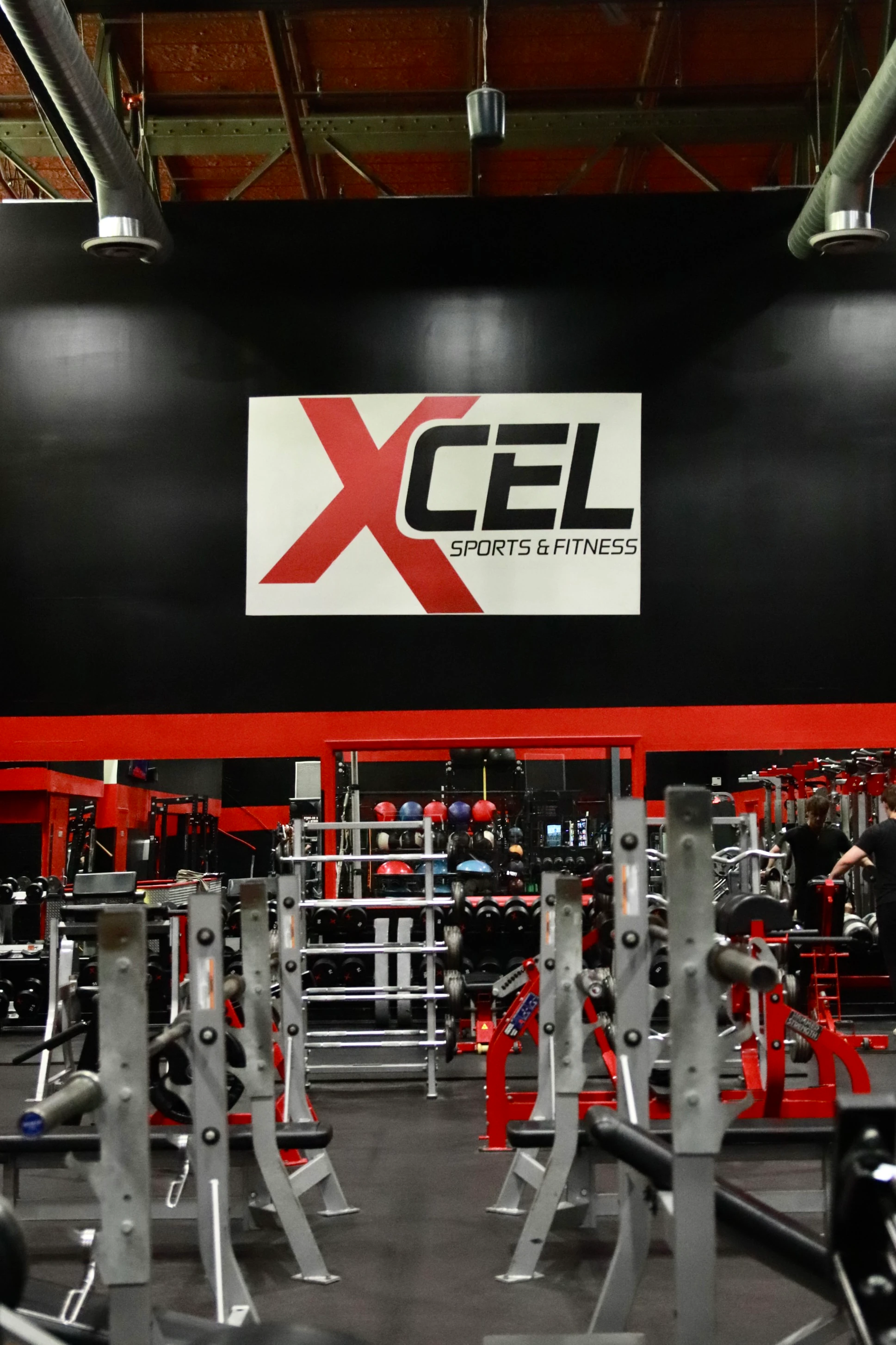 XCEL sign centered over squat racks and benches on the strength floor of the gym in Fairfield, NJ